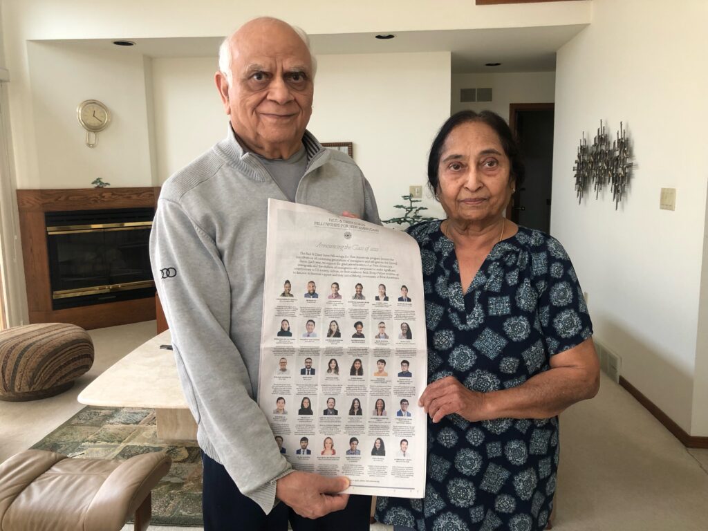 Photograph of two people inside, holding a New York Times, the man on the right is elderly and wearing a grey half-zip sweater. The woman on the right is middle-aged and wearing a blue and black patterned top. 