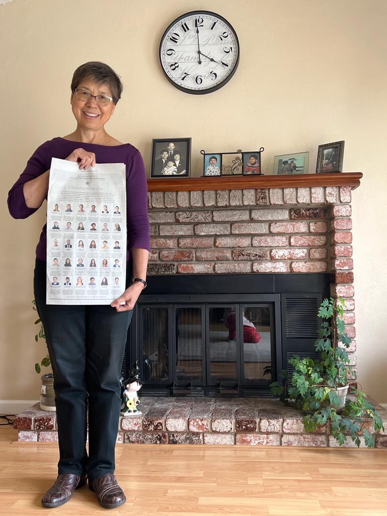 Photograph of a middle-aged woman standing in front of a fireplace, photos line the mantel, there is a clock on the wall. She is wearing a purple boat neck top and black pants, and holding the New York Times, smiling at the camera. 