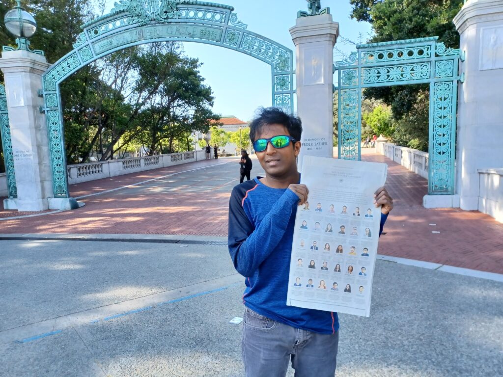 Photograph of a man in his 20s who has heritage from India standing in front of a copper green archway across a large red walkway. He is wearing a blue long sleeve top, jeans and reflective sunglasses.