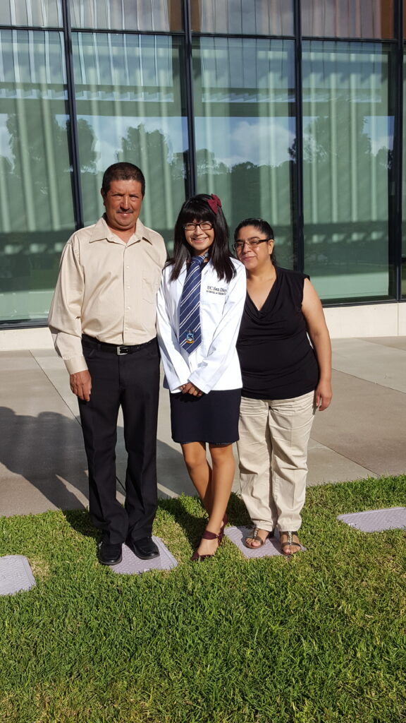 Photograph of a woman in her 20s with her parents on each side. They are standing in front of a windowed building on grass. 