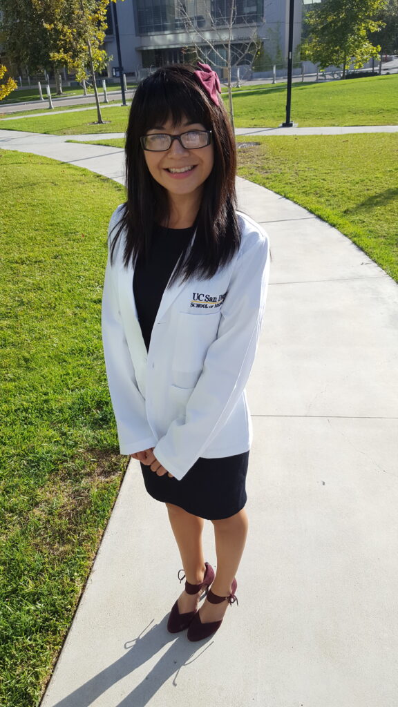 Full body photograph of a woman in her 20s who has heritage from Mexico with light skin tone and long dark brown straight hair with straight across bangs. She is wearing a doctor's white coat and "UC San Diego" can be seen embroidered on the front, underneath she has a black dress, red high heels and a red bow in her hair; she has on black rimmed rectangle glasses. She is standing on a sidewalk, with green grass on either side and a large building in the background. 