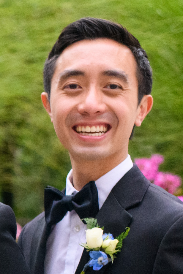 Headshot of a man in his 20s with heritage from Vietnam with light skin tone and black short hair; he is wearing a black tuxedo, with a boutonniere and is smiling at the camera.