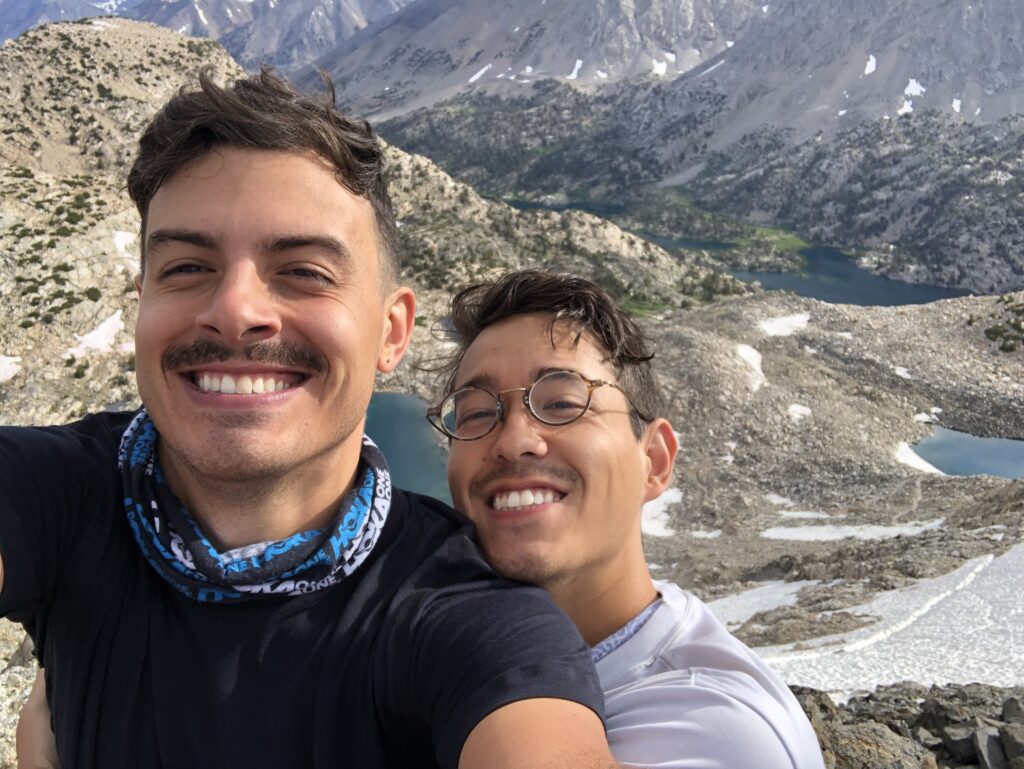 Selfie of two men in their 20s, the man not holding the camera has his arms wrapped around the other. They are both smiling at the camera and there is a vast landscape behind them with mountains and pools of water. 