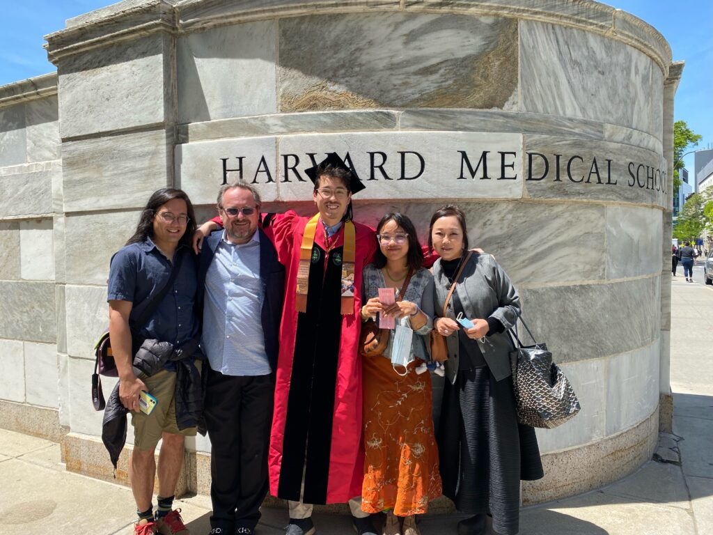 Photograph of five people, the man in the center is wearing graduation robes and hat. They are standing in front of a stone wall that says "Harvard Medical School". 