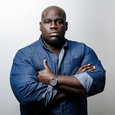 Headshot of a man in his 30s with heritage from Jamaica he has dark skin tone, he is bald. He is wearing a denim button up shirt and a watch on his right wrist. His arms are crossed, and he looks directly at the camera.