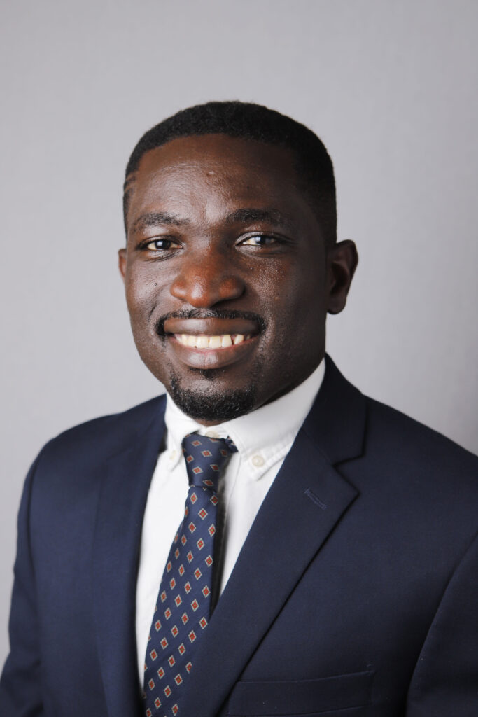 Headshot of a man in his 20s who has heritage from Nigeria with dark skin tone, black buzzed hair, and a trimmed mustache/ goatee. He is wearing a navy suit, white button up shirt and navy tie. He is smiling at the camera.