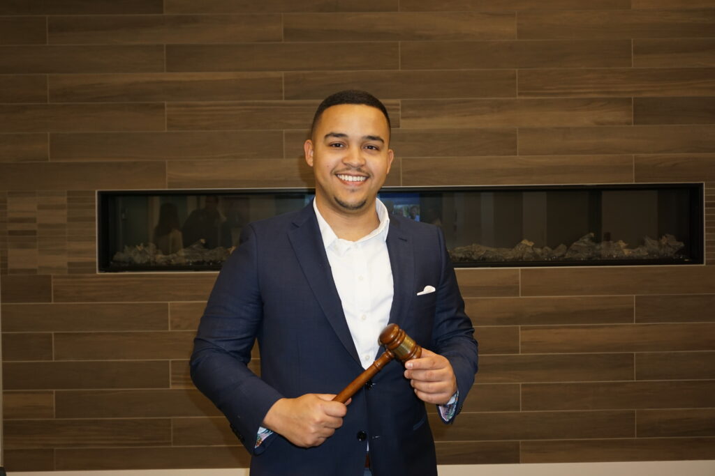 Half body photograph of a man in his 20s who has heritage from Dominican Republic with medium skin tone, buzzed black hair and a trim goatee, mustache. He is wearing a black suit with a white pocket square and white button up shirt. He is standing in front of a wood wall, holding a gavel. 