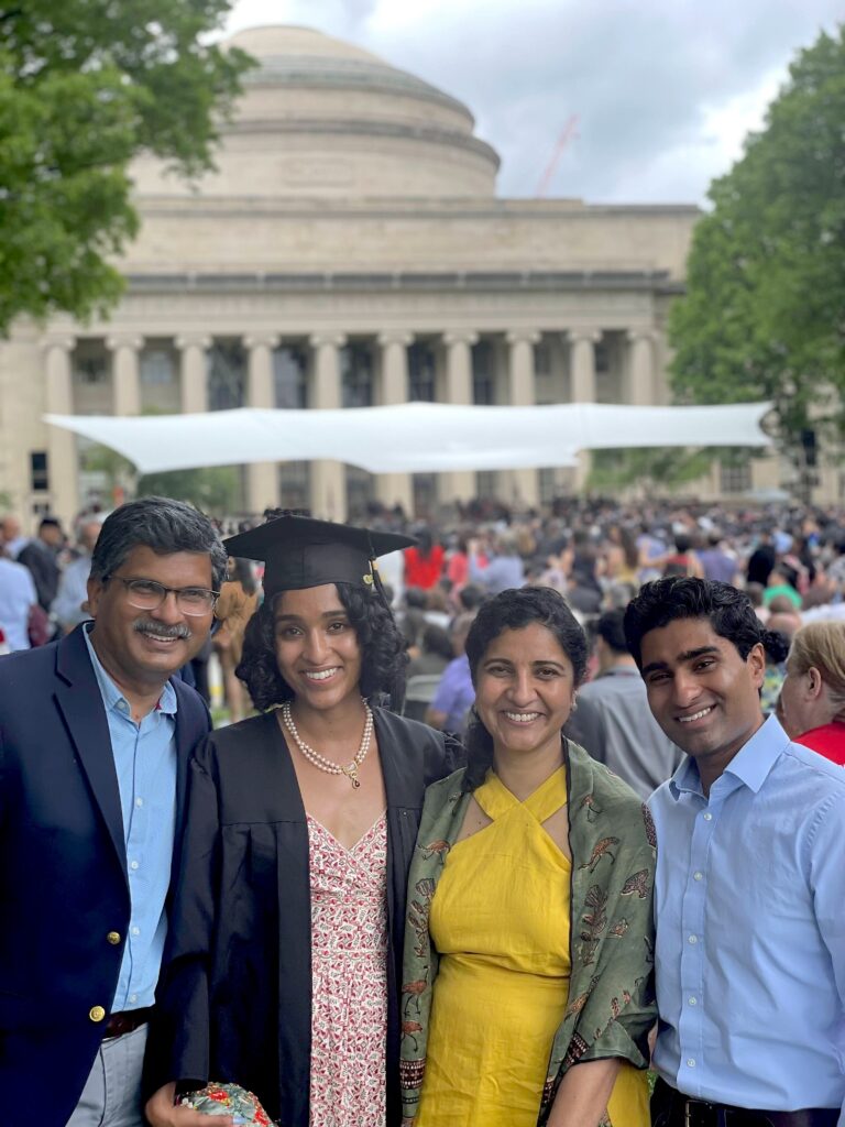 Photograph of four adults standing together, posing and smiling for the camera. There is a large domed and columned building behind them and a large crowd can be seen between them and the building. The person second from the left is wearing a graduation cap and gown. 