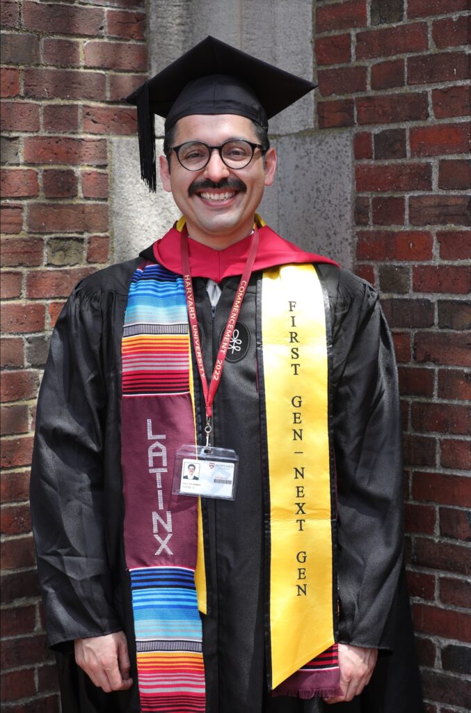 Half body photograph of a man in his 20s who has heritage from Mexico with light skin tone, black combed back hair, and a mustache. He is wearing black graduation robes and hat with two banners down his torso - one says "Latinx" with many colors, the second says "First Gen / Next Gen" on yellow and an ID badge on a lanyard. He has large round black rimmed glasses on. He is smiling at the camera. 