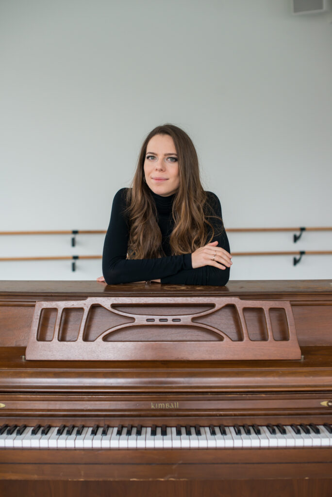 Photograph of a woman in her 20s who has heritage from Russia with light skin tone and long wavy light brown hair parted in the middle. She is wearing a black long sleeve turtleneck and has a few silver rings on. She is standing behind a piano, resting her crossed arms on top of the piano, smiling at the camera.