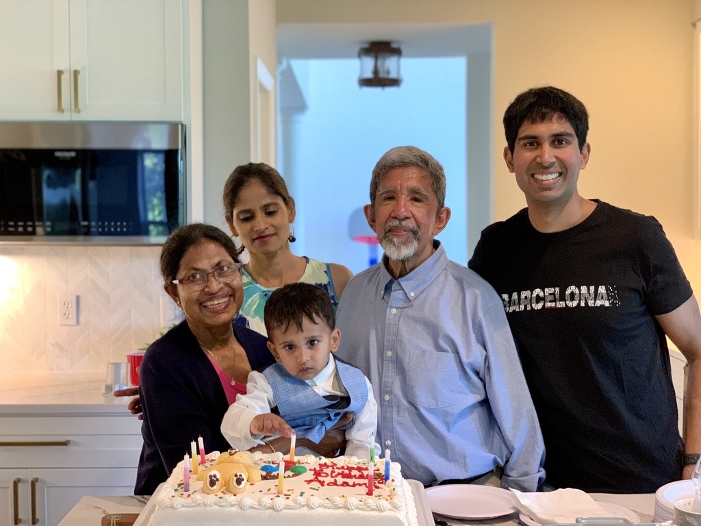 Photograph of a family standing in a kitchen, there are four adults and one child, on the counter in front of them there is a large sheet cake decorated with a teddy bear and candles and it says "Happy Birthday"