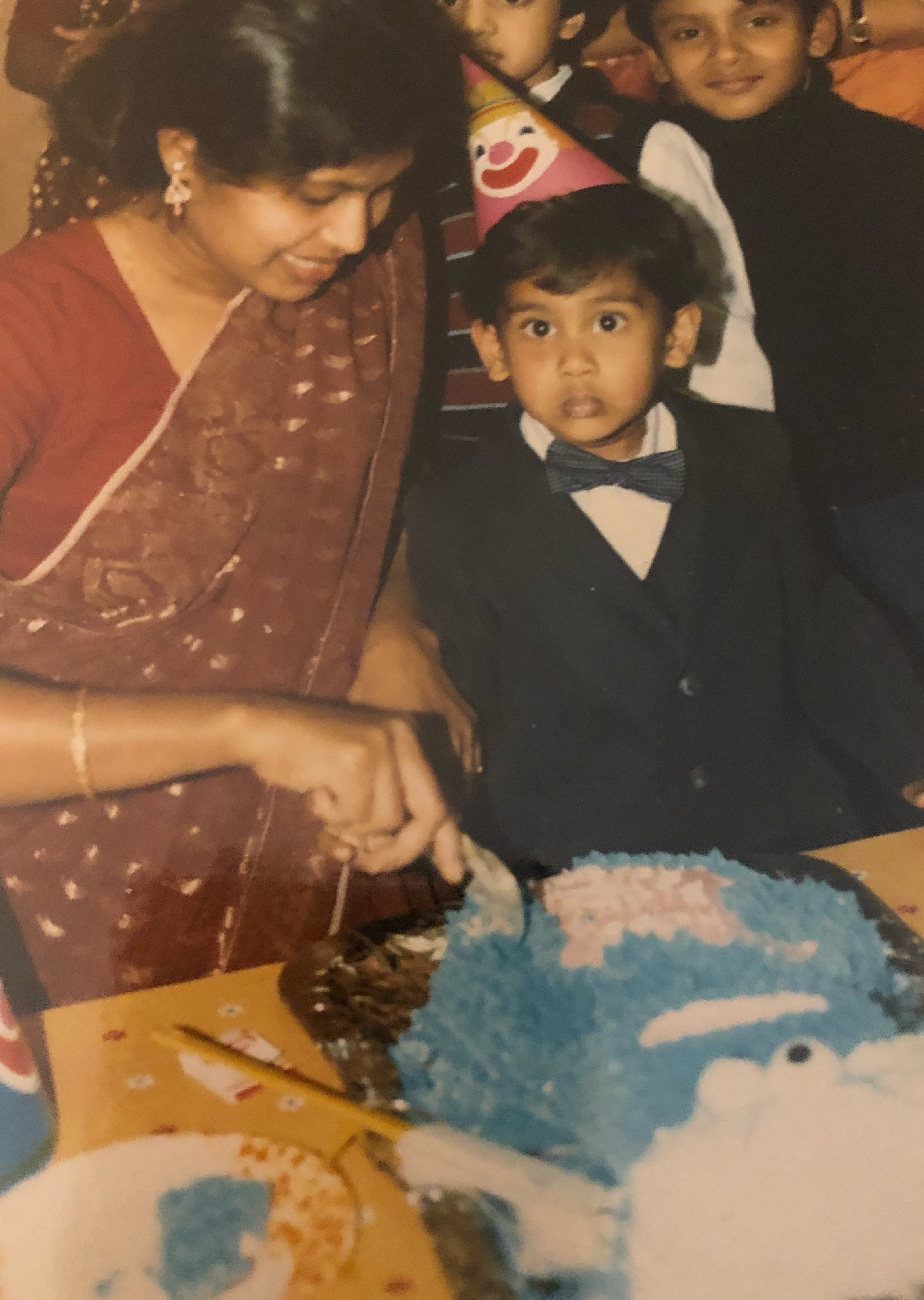 Old photograph of an adult woman dressed in a red and gold saari and cutting a cake that is decorated to look like Cookie Monster. A young boy in a tuxedo stands next to her looking at the camera and wearing a birthday hat with a clown on it.