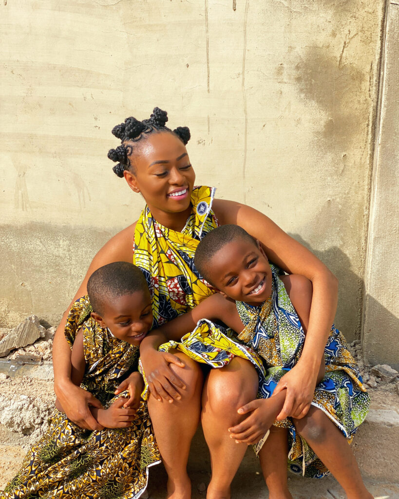 Photograph of a woman in her 20s who has heritage from Nigeria with medium skin tone and black hair tied into Bantu knots. She is wearing a multi-colored sleeveless dress and hugging two young boys sitting on either side of her. 