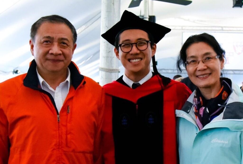 Photograph of three people, the man in the middle is in his 20s who has heritage from China with light skin tone; he is wearing graduation robe, red hood and a cap. His parents are on each side, they are all smiling at the camera.  