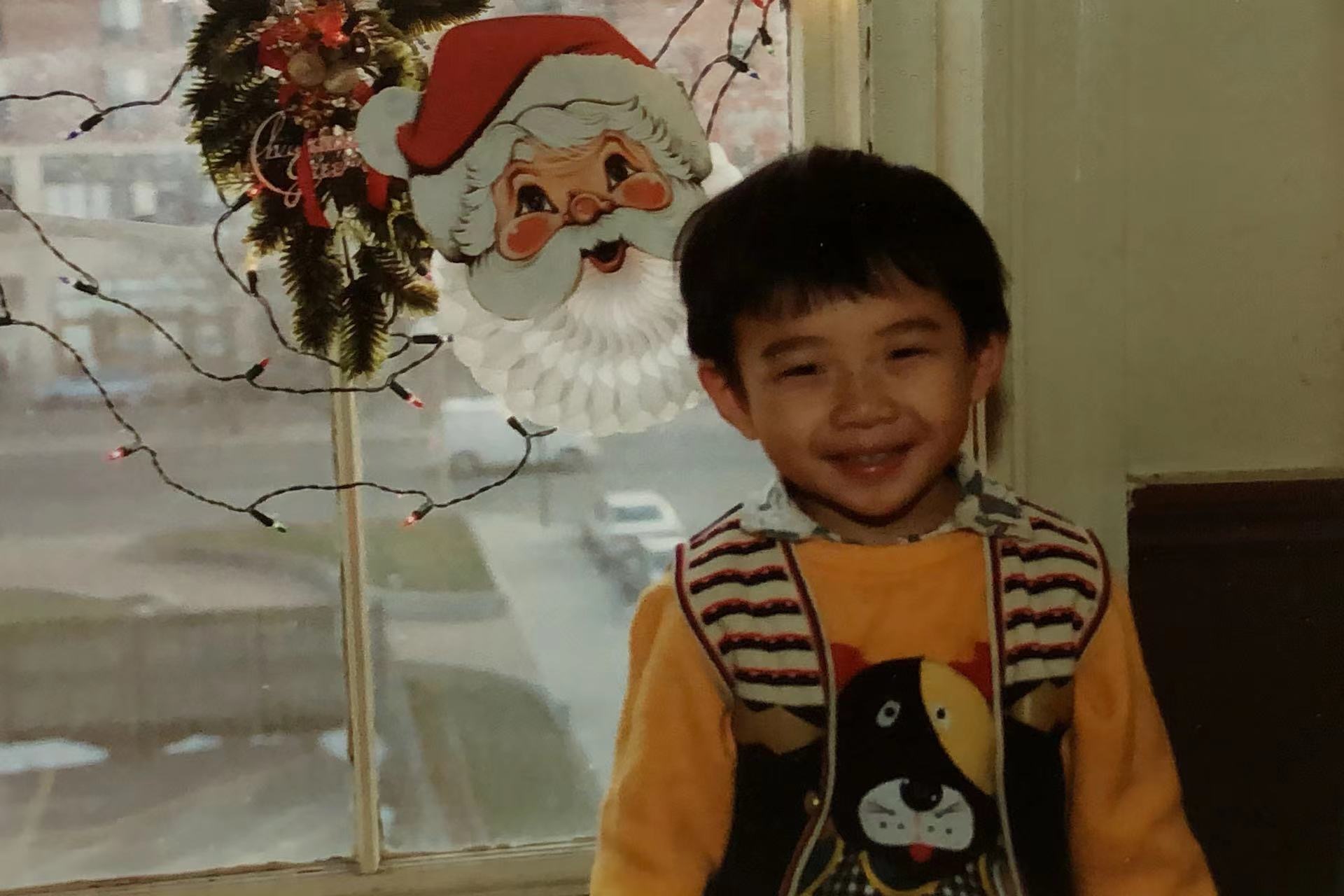 Old photograph of a child, maybe 4, wearing a bright yellow sweatshirt with a cartoon animal, button up shirt under and a multi-colored vest. He is standing next to a few Christmas decorations in a window. 