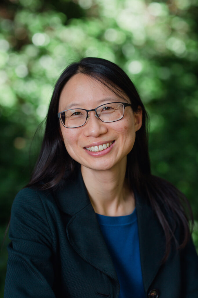 Headshot of a woman in her 20s who has heritage from Taiwan with light skin tone and long straight black hair parted in the middle. She is wearing a dark teal blazer, blue blouse and black thin rimmed rectangle glasses. She is smiling at the camera. 