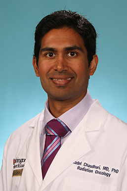 Headshot of a man in his 30s with heritage from Bangladesh and India, who has medium skin tone and black hair. He is wearing a white doctor's coat with his name, department and hospital embroidered, light purple button up shirt and marron tie. He is smiling at the camera. 