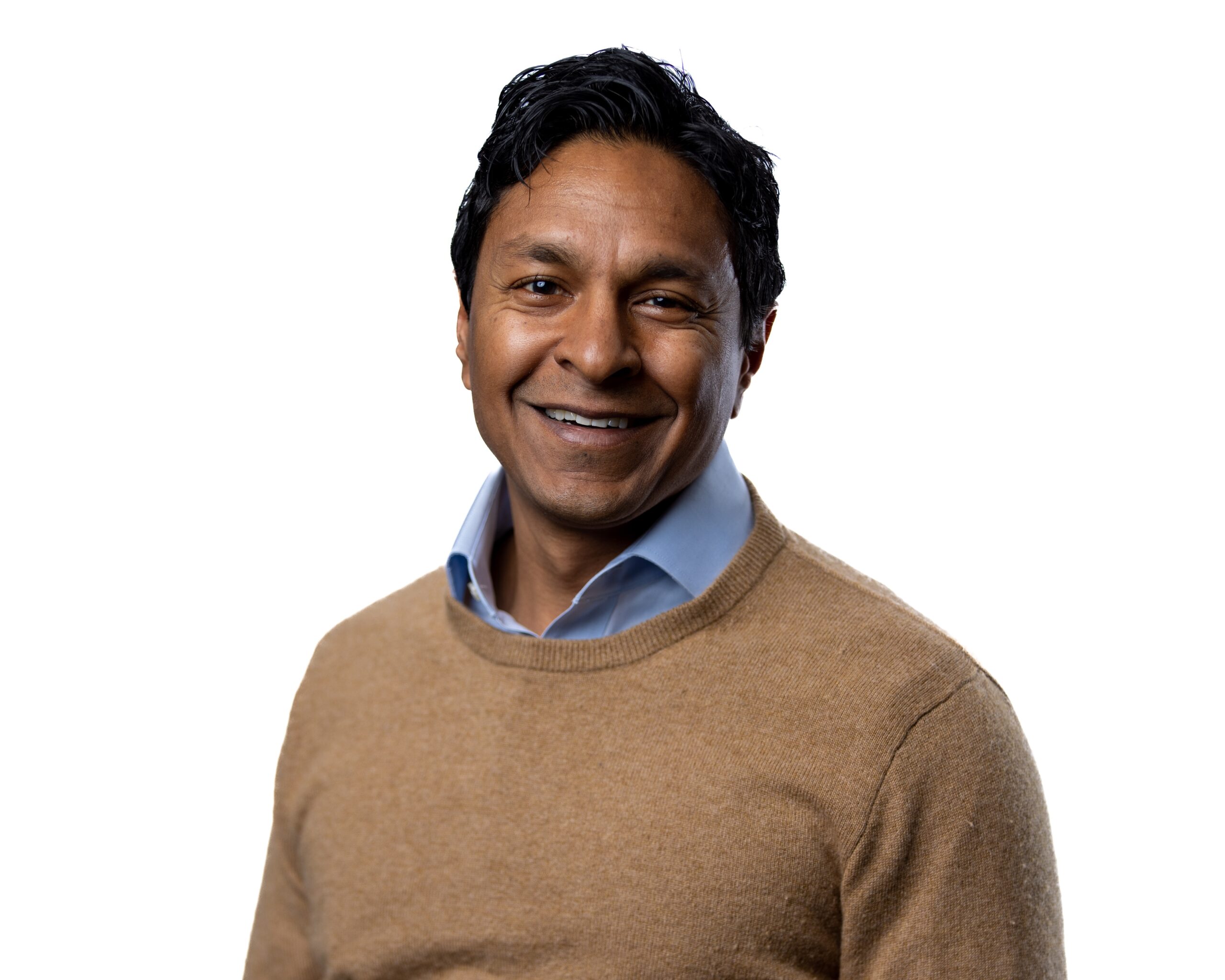 Headshot of a man in his 40s who has heritage from India with medium-dark skin tone and black hair. He is wearing a blue button up shirt with a tan crew neck sweater on top. He is smiling at the camera.