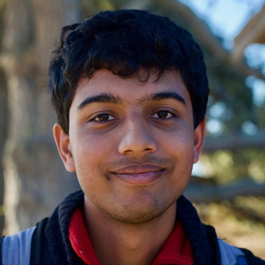 Headshot of a man in his 20s who has heritage from India, with medium skin tone and black hair. He is smiling at the camera.