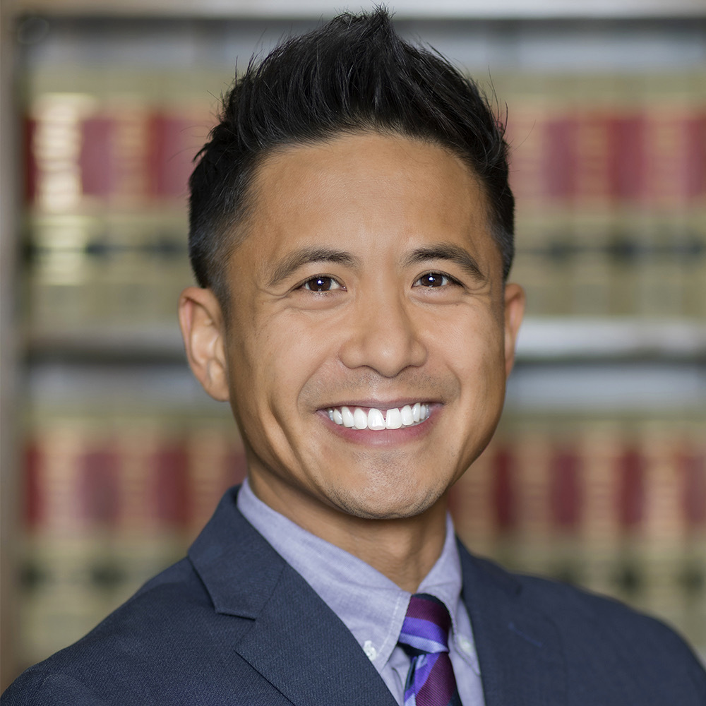 Headshot of a man in his 40s who has heritage from the Philippines with light-medium skin tone and black crew cut hair. He is wearing a navy suit, light purple button up and maroon and purple tie. He is smiling at the camera.