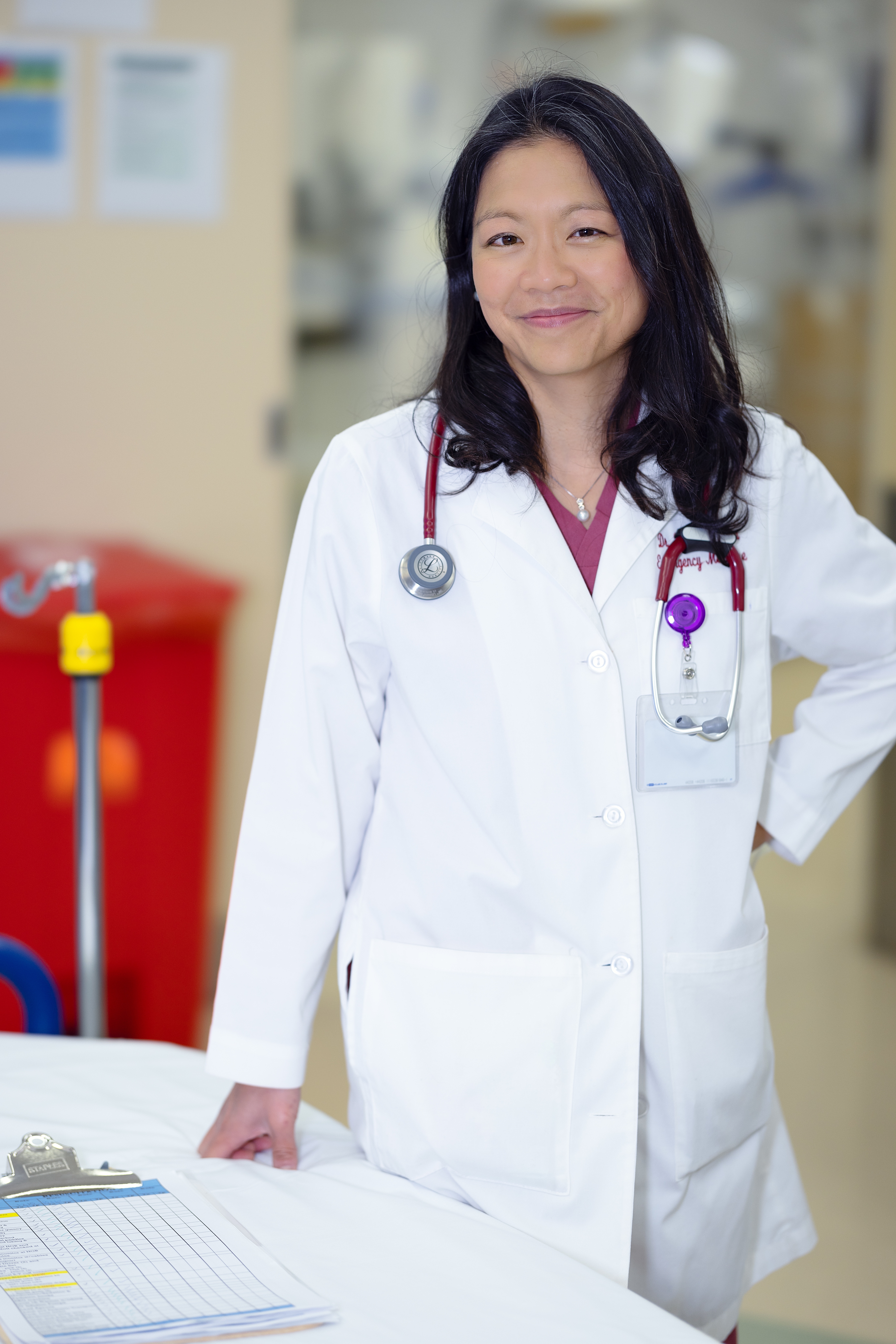 Photograph of a woman standing at a table, her right-hand rests on the table, her left hand is on her hip. She is in her 40s and has heritage from China, with light skin tone and just passed shoulder length black curled hair. She is wearing a doctor's white coat with a red shirt under, she has a stethoscope around her neck and a badge on her left lapel. She is smiling at the camera. 