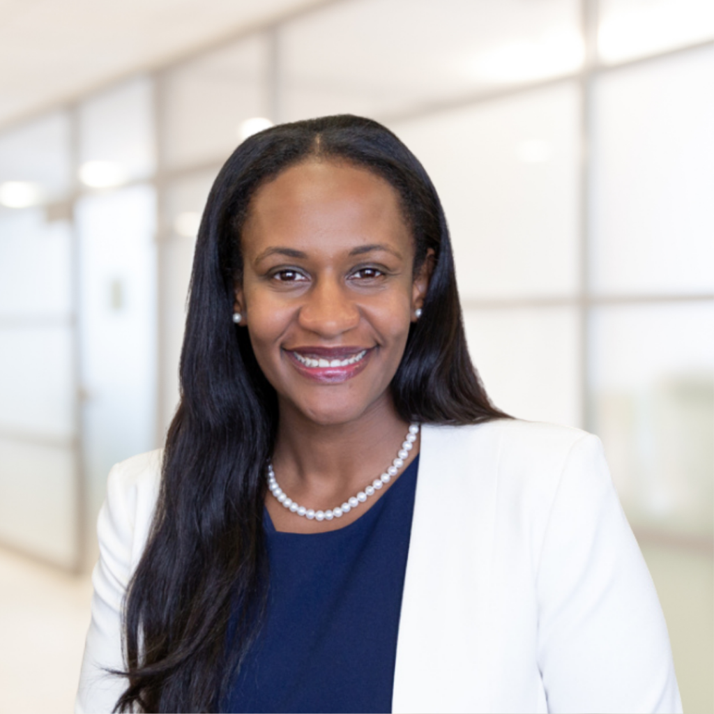 Headshot of a woman in her 30s who has heritage from Ethiopia with dark-medium skin tone and long black wavy hair parted in the middle. She is wearing a white jacket with a blue blouse, pearl necklace and pearl studs. She is looking at the camera and smiling. 