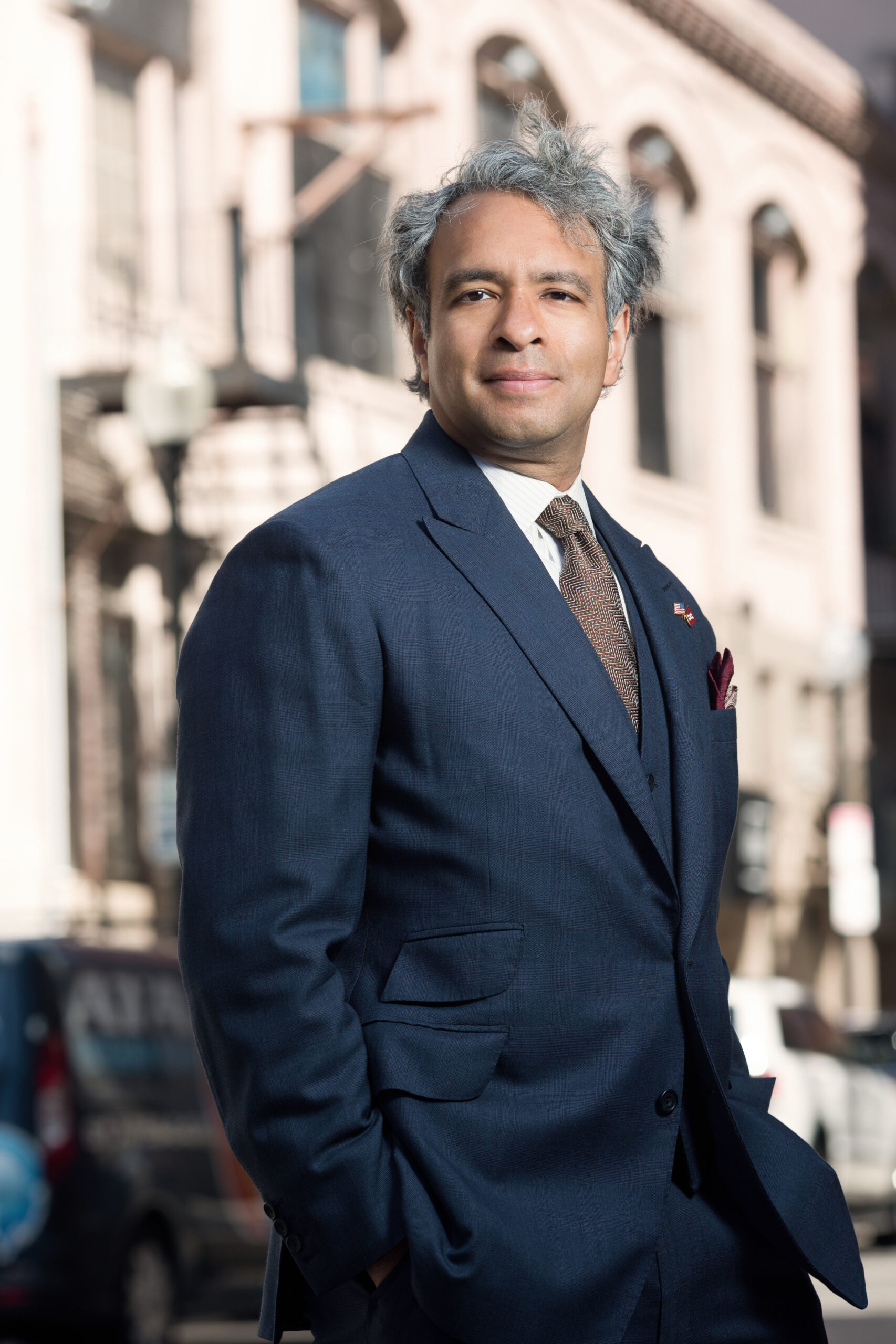 Headshot of a man in his 40s who has heritage from India with light-medium skin tone and salt and pepper short hair. He is wearing a navy suit, white button up shirt and brown tie. He is standing with his hands in his pockets looking at the camera. 