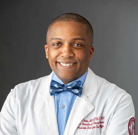 Headshot of a man in his 40s who her heritage from Haiti with medium skin tone and buzzed black hair. He is wearing a doctor's white coat, with his name embroidered in red on his left side, under the coat he has a light blue button up and blue bow tie. He is smiling at the camera.