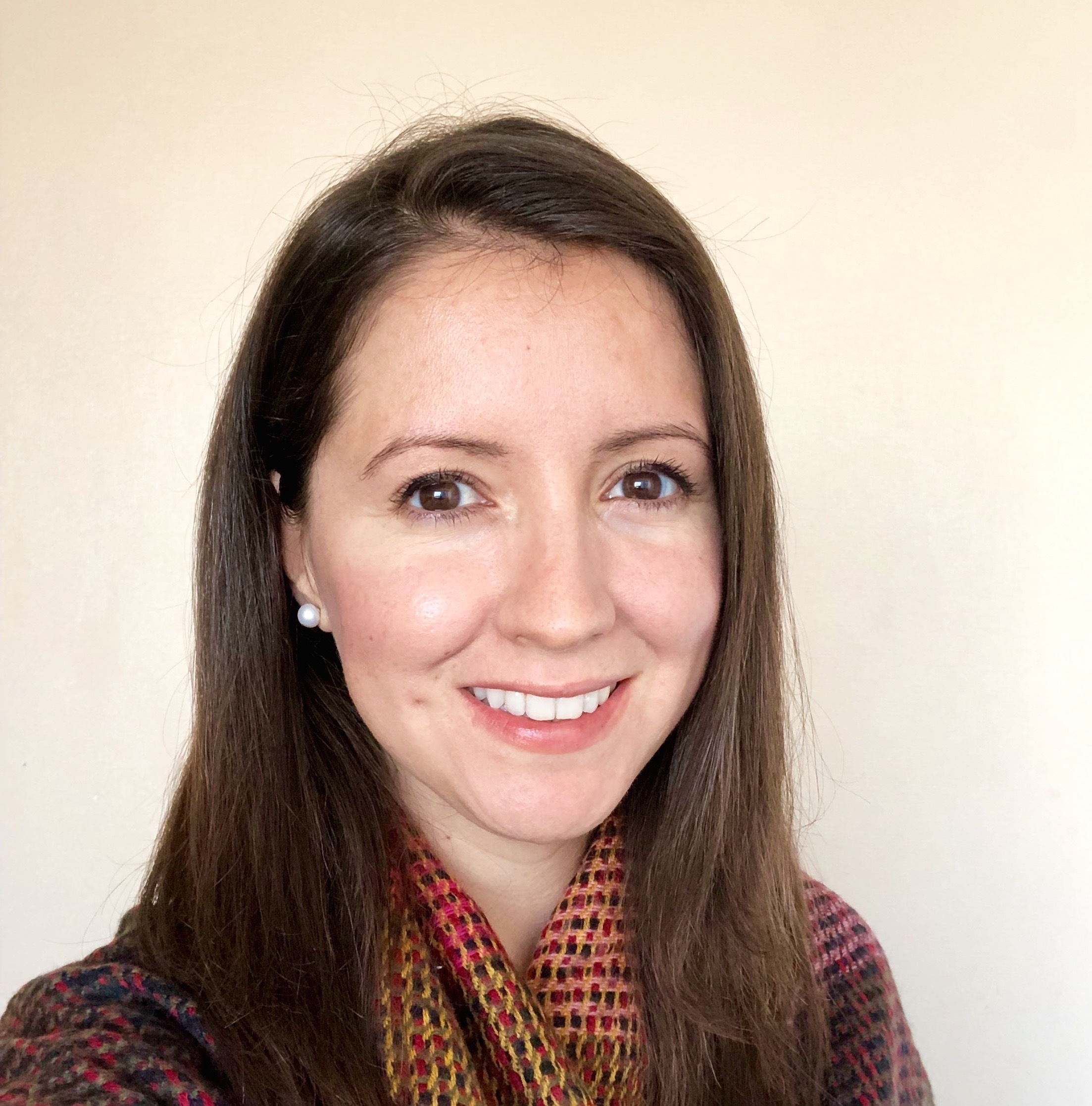 Headshot of a woman in her 20s who has heritage from Mexico with light skin tone and long, straight brown hair. She is wearing a multi-colored sweater and pearl stud earrings. She is smiling at the camera.