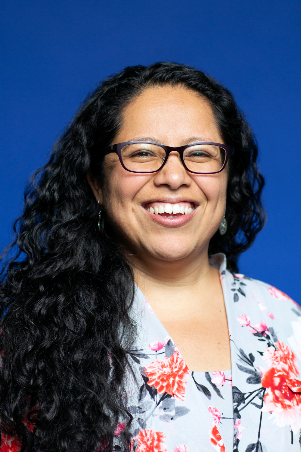 Headshot of a woman in her 40s, who has heritage from Mexico, with light skin tone and long curly black hair, pulled over her right shoulder. She is wearing a matching white with red, black and pink flower shirt and sweater set, and black framed rectangle glasses. She is smiling at the camera.