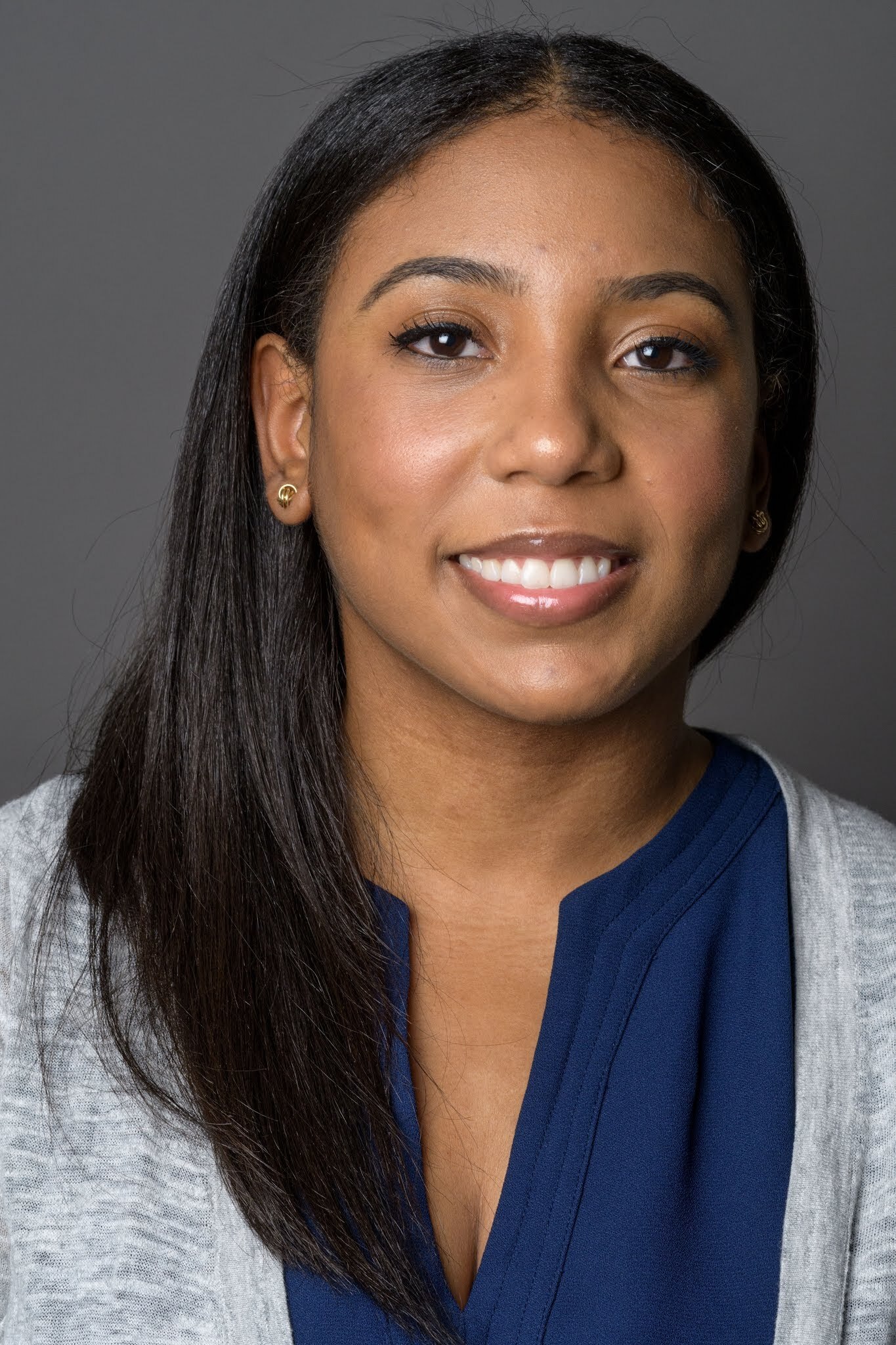 Headshot of a woman in her 20s with heritage from Dominican Republic, she has medium skin tone and straight, long black hair. She is wearing a light grey sweater, blue shirt and smiling at the camera.