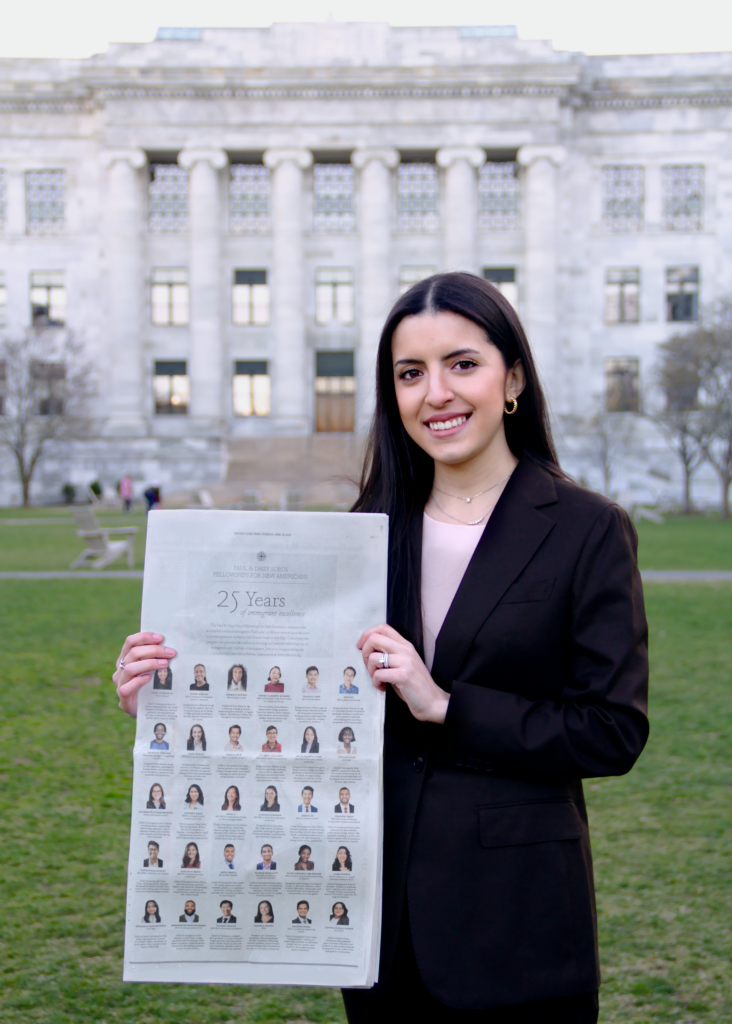 Photograph of a woman in her 20s who has heritage from Colombia with light skin tone and long dark brown straight hair. She is standing on a lawn with a large white stone building in the background. 