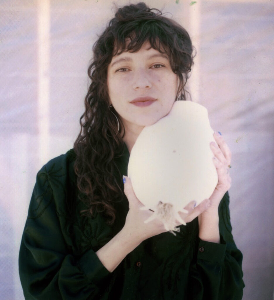 Photograph of a woman in her 20s who has heritage from Colombia with light skin tone and long dark brown curly hair. She is wearing a dark green long sleeve blouse and silver dangly earrings. She is holding a white sculpture with both hands, and gently resting her chin on it.