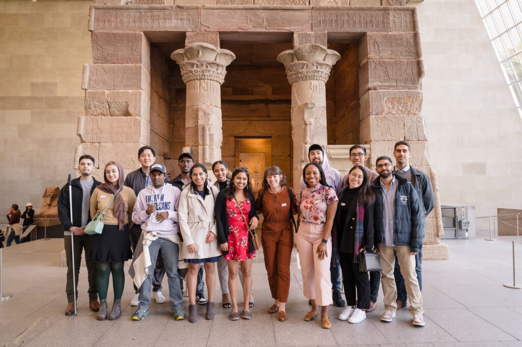 Photograph of a group of people standing in front of the Temple of Dendur at the Metropolitan museum. This is a temple that has a reddish color, from Egypt that now lives inside a room with a wall of windows.