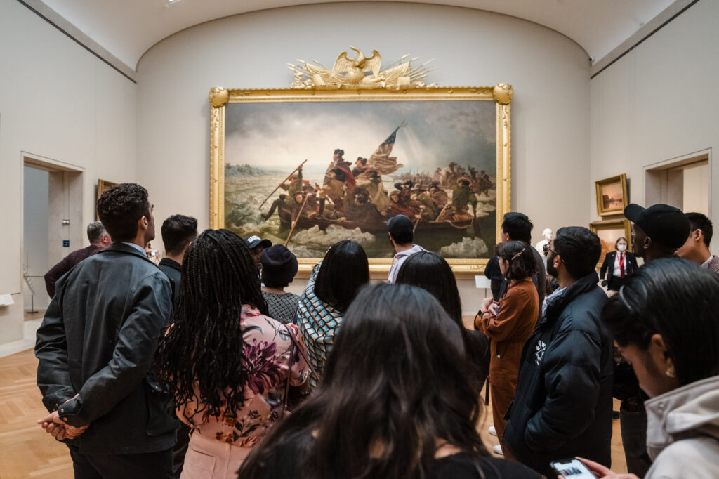 Photograph of a group of people standing in a museum gallery viewing a large painting. 