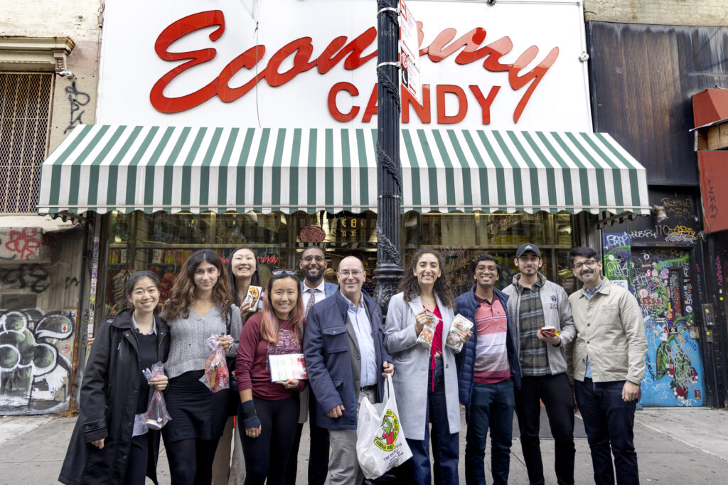 Photograph of a group of people posing in front of the candy store "Economy Candy", which appears in red lettering on a white background, in front of the sign is a white and green striped awning. 