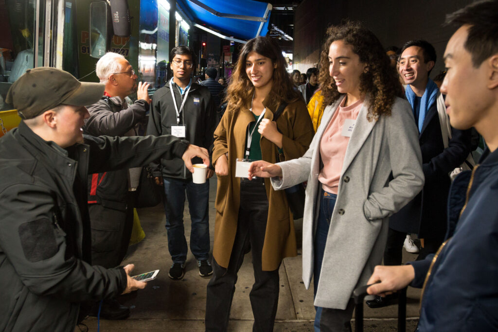 Photograph of a group of people standing on a street under awnings. A person is handing out small white cups.