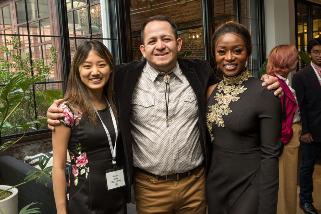 Photograph of three people in their 20s with varying heritages, dressed in cocktail attire, with their arms around each other looking at the camera and smiling. 