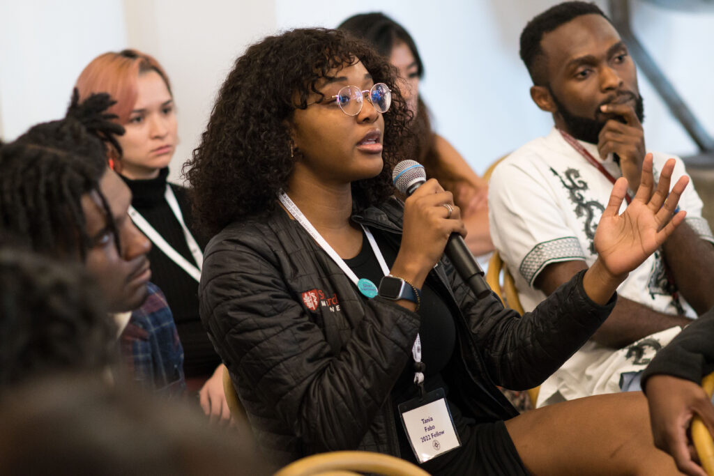 Photograph of a woman in her 20s holding a microphone and speaking. There are people sitting around here, everyone looking in the same direction and with a serious look on their faces. 