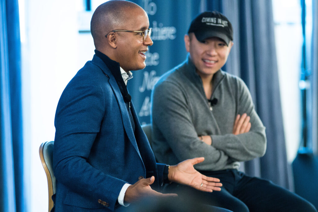 Photograph of two men sitting, they appear to be on a stage with a blue curtain behind them. They are looking off camera and the man closer to the camera is smiling and gesturing with his hands. 