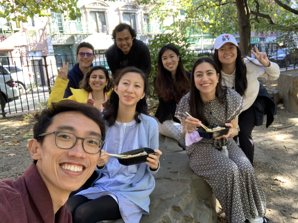 Photograph of a group of people in their 20s siting in a park, some of them are eating.