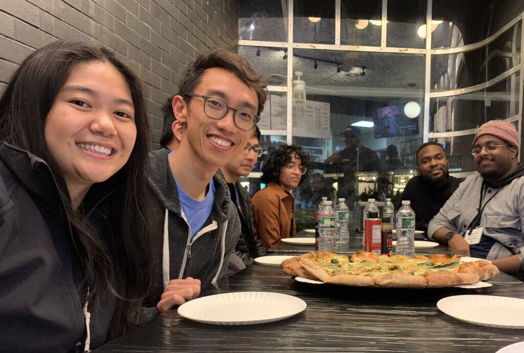 Photograph of a group of people in their 20s who have a variety of heritages, sitting around a table, pizza and drinks are in the middle of the table.