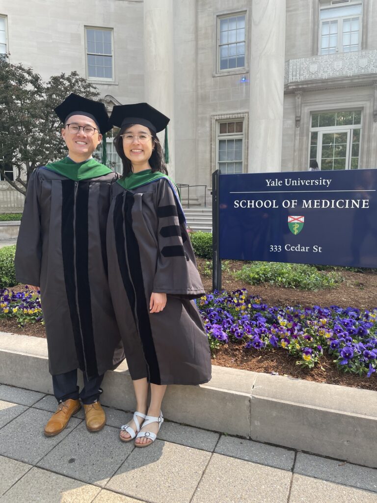 Photograph of a man in his 20s who has heritage from Taiwan with light skin tone, a woman stands next to him. They are both wearing graduation robes and caps. There is a sign behind them that says "Yale University School of Medicine" purple pansies line the ground.