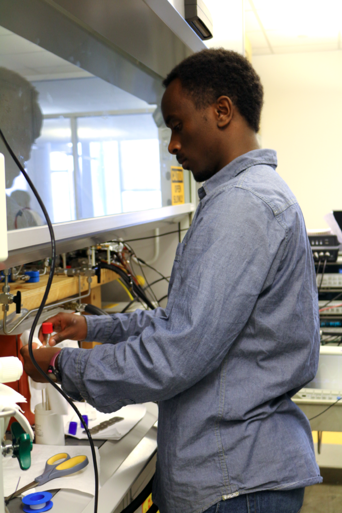 Photograph of a man in his 20s who has heritage from Ethiopia with dark skin tone and short black hair. He is wearing a denim blue button up and standing in a lab, not looking at the camera. 