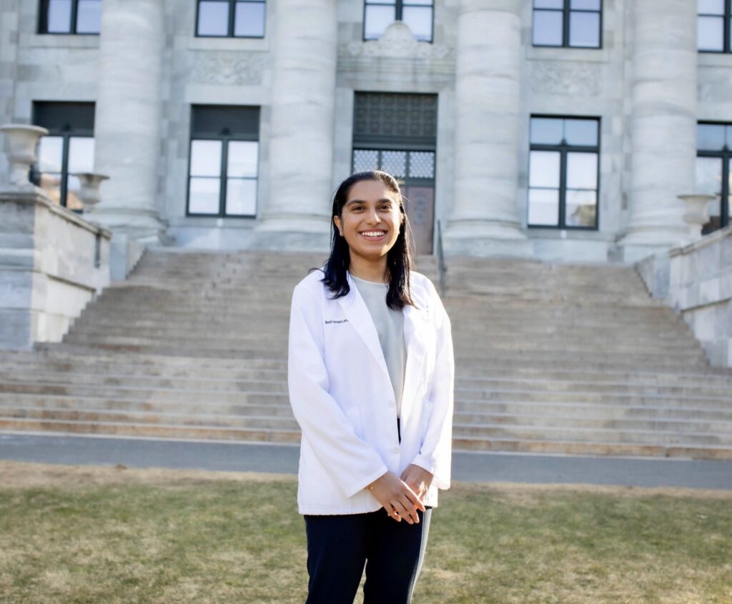 Photograph of a woman in her 20s who has heritage from India with light-medium skin tone and long black hair. She is wearing a doctor's white coat, light blue blouse and black pants. She is holding her hands together and smiling at the camera. She is standing in front of a large white stone staircase and building.