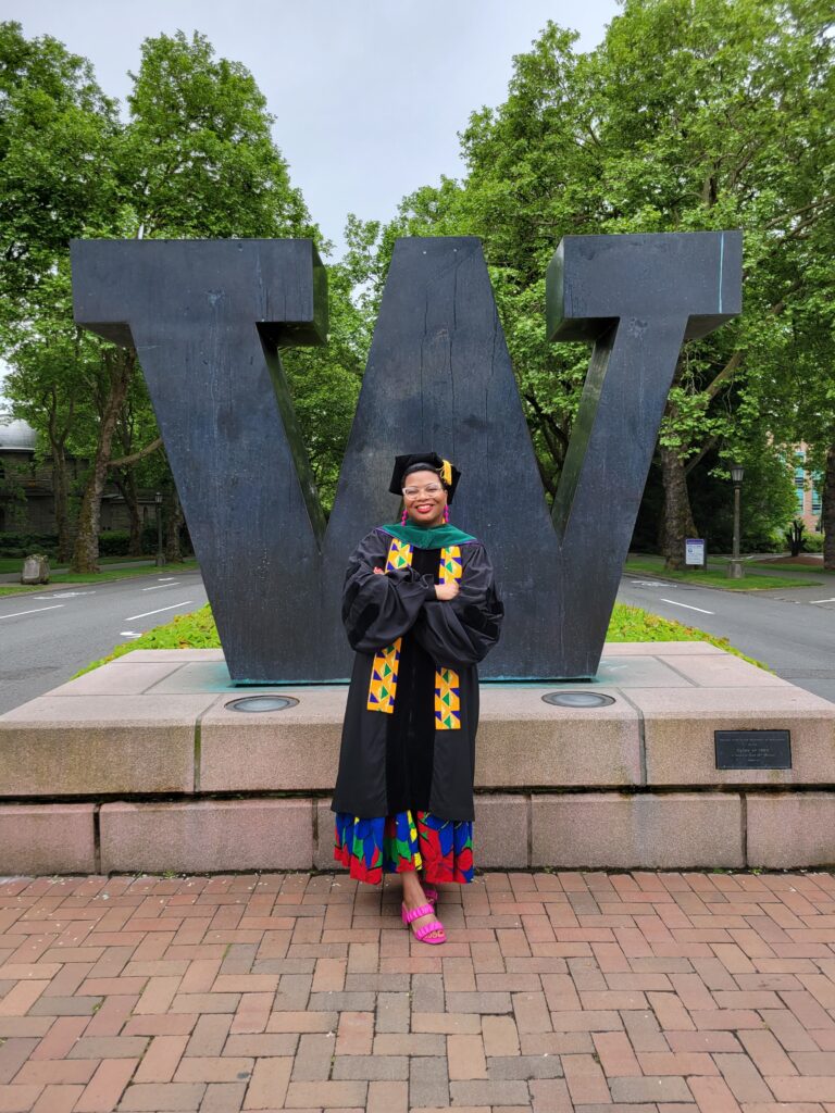 Photograph of a woman in her 20s who has heritage from Cameroon of Congolese with medium-dark skin tone. She is wearing black graduation robes, a green hood, a brightly colored dress can be seen. She is standing in front of a large "W" sculpture, crossing her arms and smiling.