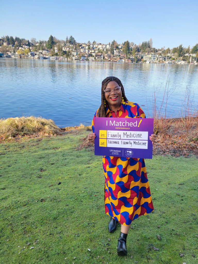 Photograph of a woman in her 20s who has heritage from Cameroon of Congolese with medium-dark skin tone and black braided black hair. She is wearing a brightly colored patterned dress and holding a sign that says "I matched! in Family Medicine at Tacoma Family Medicine". She is standing on a green lawn, with a body of water behind her and buildings can be seen across the water. 