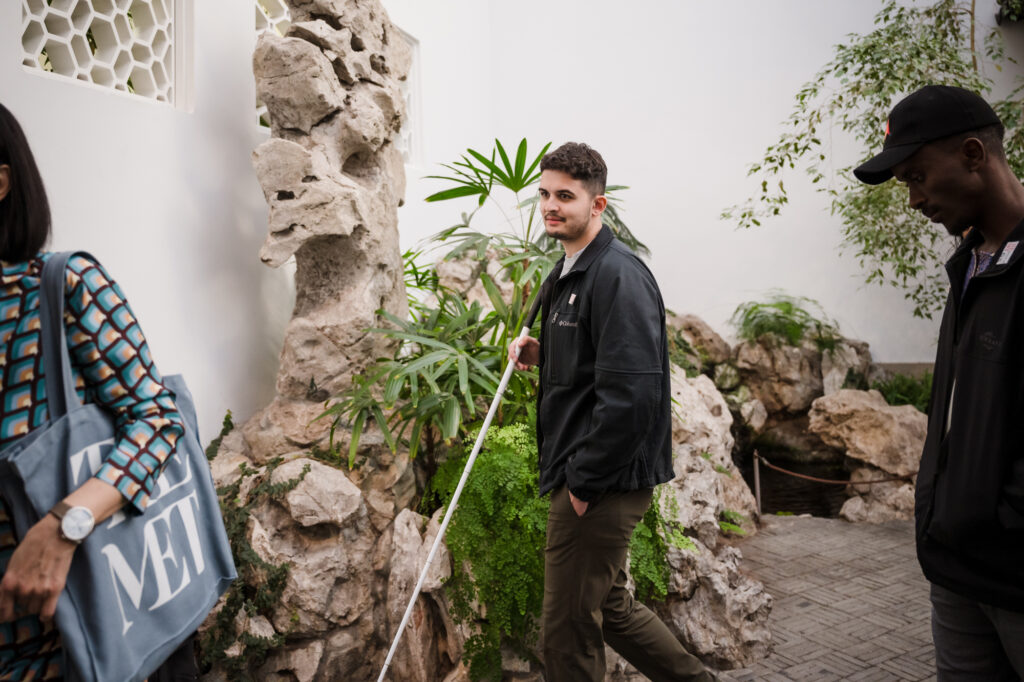 Photograph of a man in his 20s who has heritage from Pakistan with light skin tone, crew cut dark brown hair and short beard. He is wearing a black jacket and brown pants. He holds a guide cane and stands next to rock formations and plants.