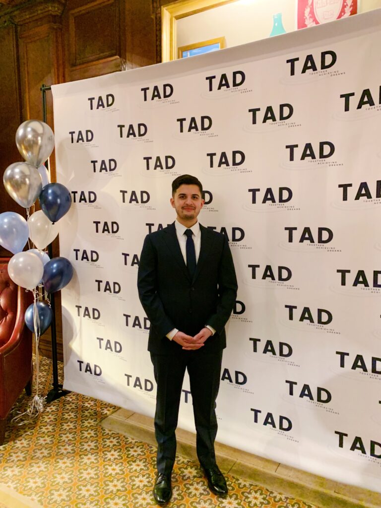 Photograph of a man in his 20s who has heritage from Pakistan with light skin tone, crew cut dark brown hair and short beard. He is standing in front of a white step & repeat with "TAD" in black. He is wearing a black suit and smiling at the camera.