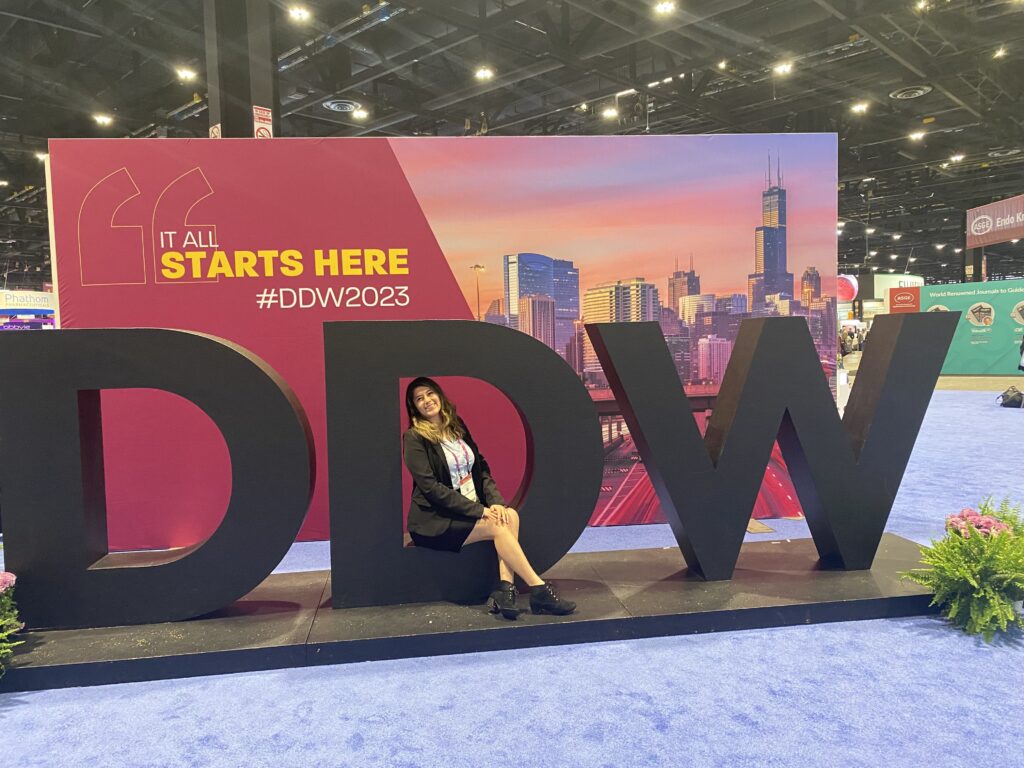 Photograph of a woman wearing a black blazer, black skirt, white blouse and black shoes, she is sitting inside a large letter "D", it is the middle D of the initials "DDW" with a sign in the background stated "It all starts here #DDW2023".